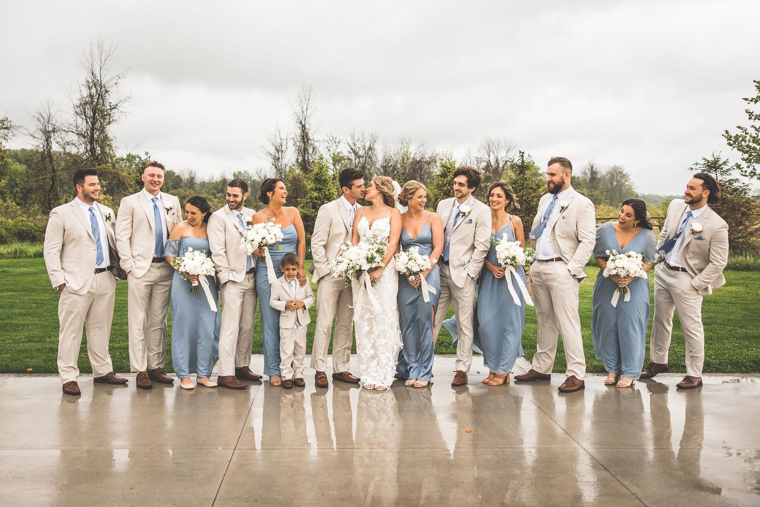 Wedding party lined up on wet concrete with bridesmaids in blue and groomsmen in beige.