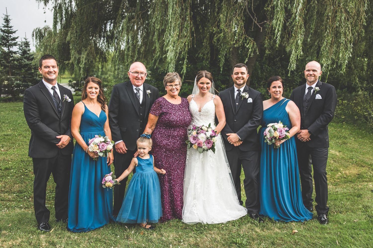 Wedding party in blue dresses and black suits posing under weeping willow trees.