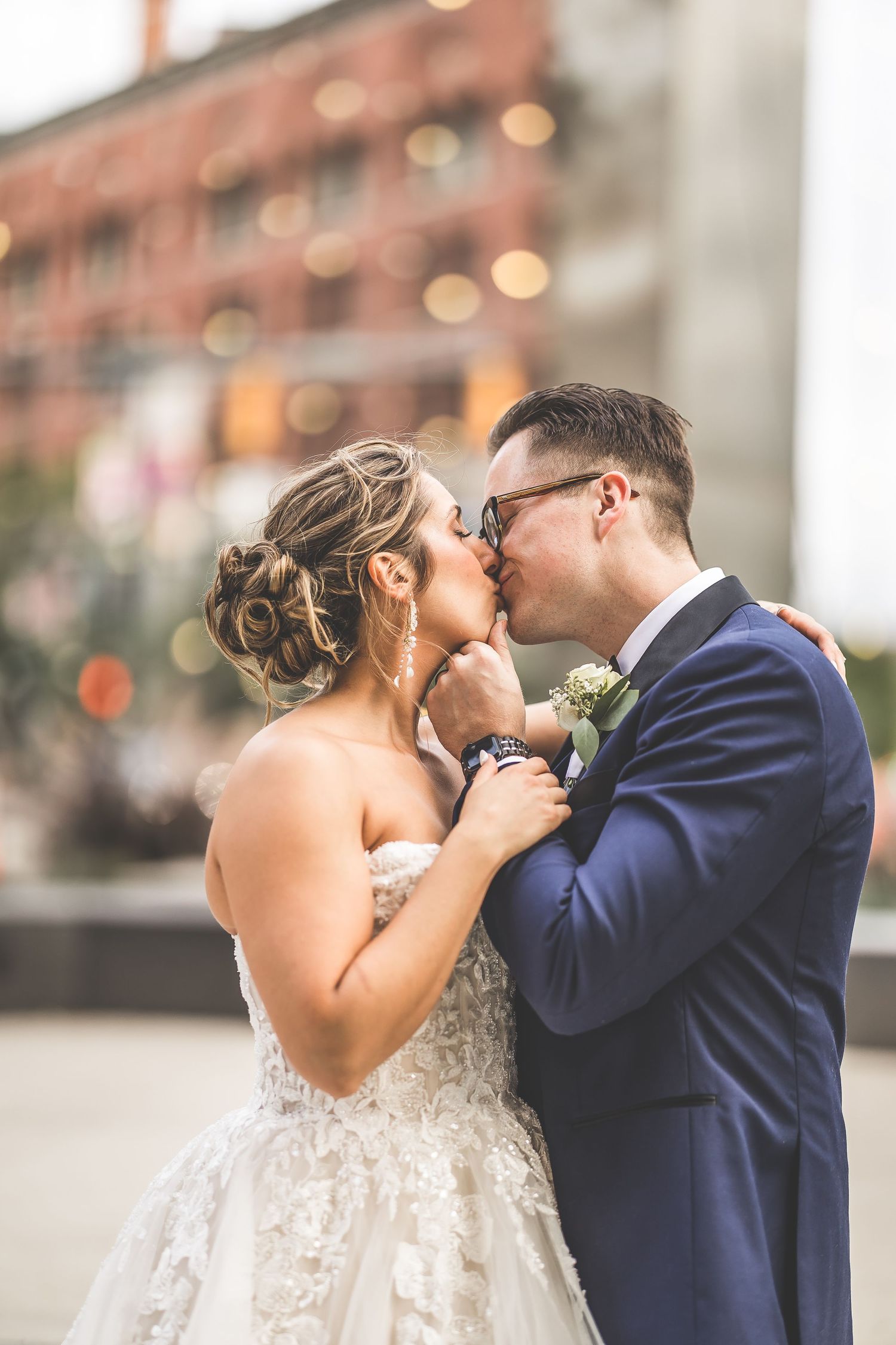 A romantic wedding kiss between a couple, with the bride in lace and groom in navy suit against an urban backdrop.