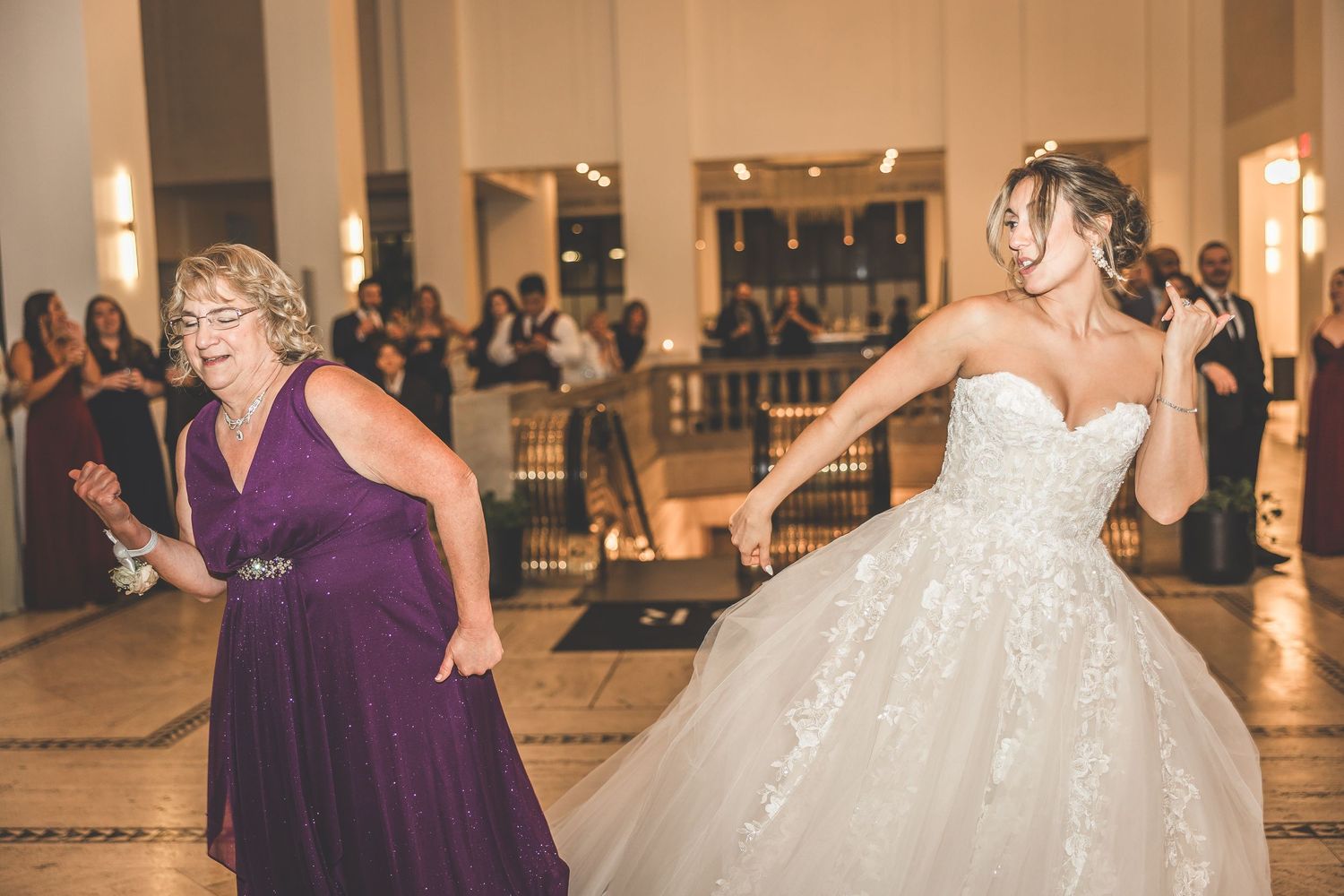 Bride in sparkly ballgown and guest in purple dress dancing energetically at wedding celebration.