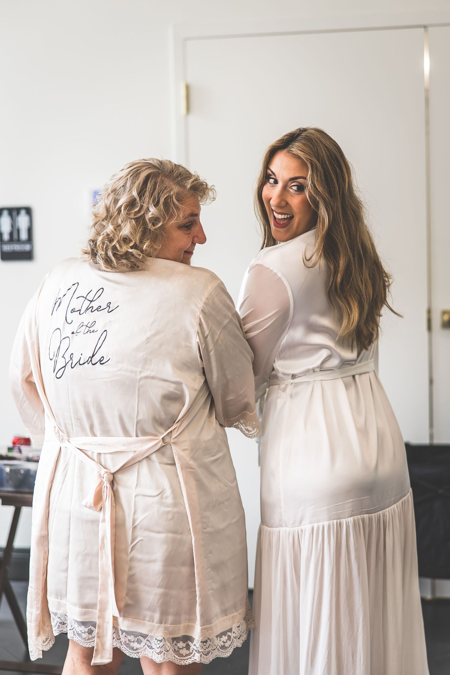Two people wearing matching white satin robes with lace trim during wedding preparations.