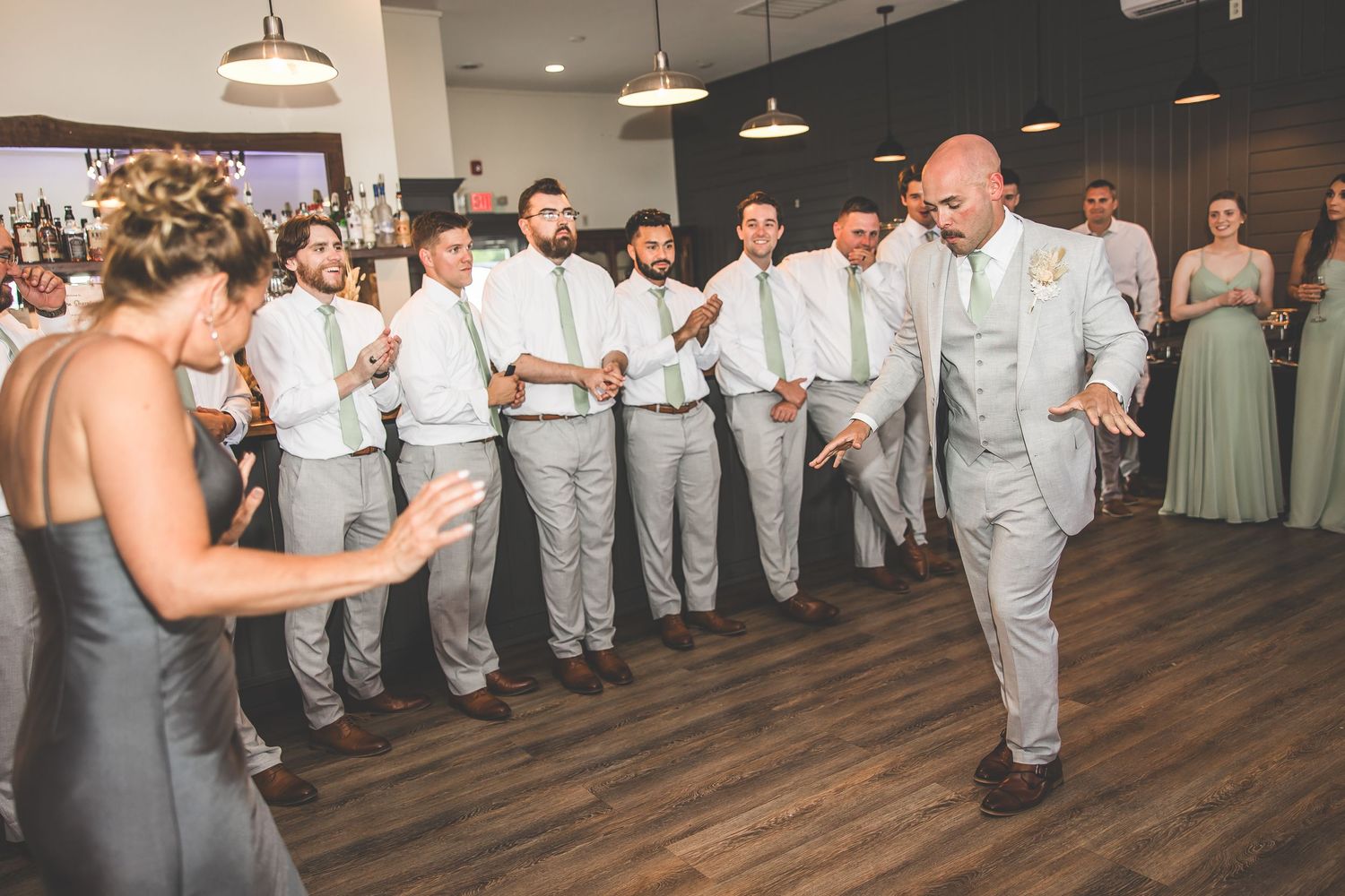 Wedding party members in gray suits and bridesmaids in sage dresses line up on wooden dance floor.