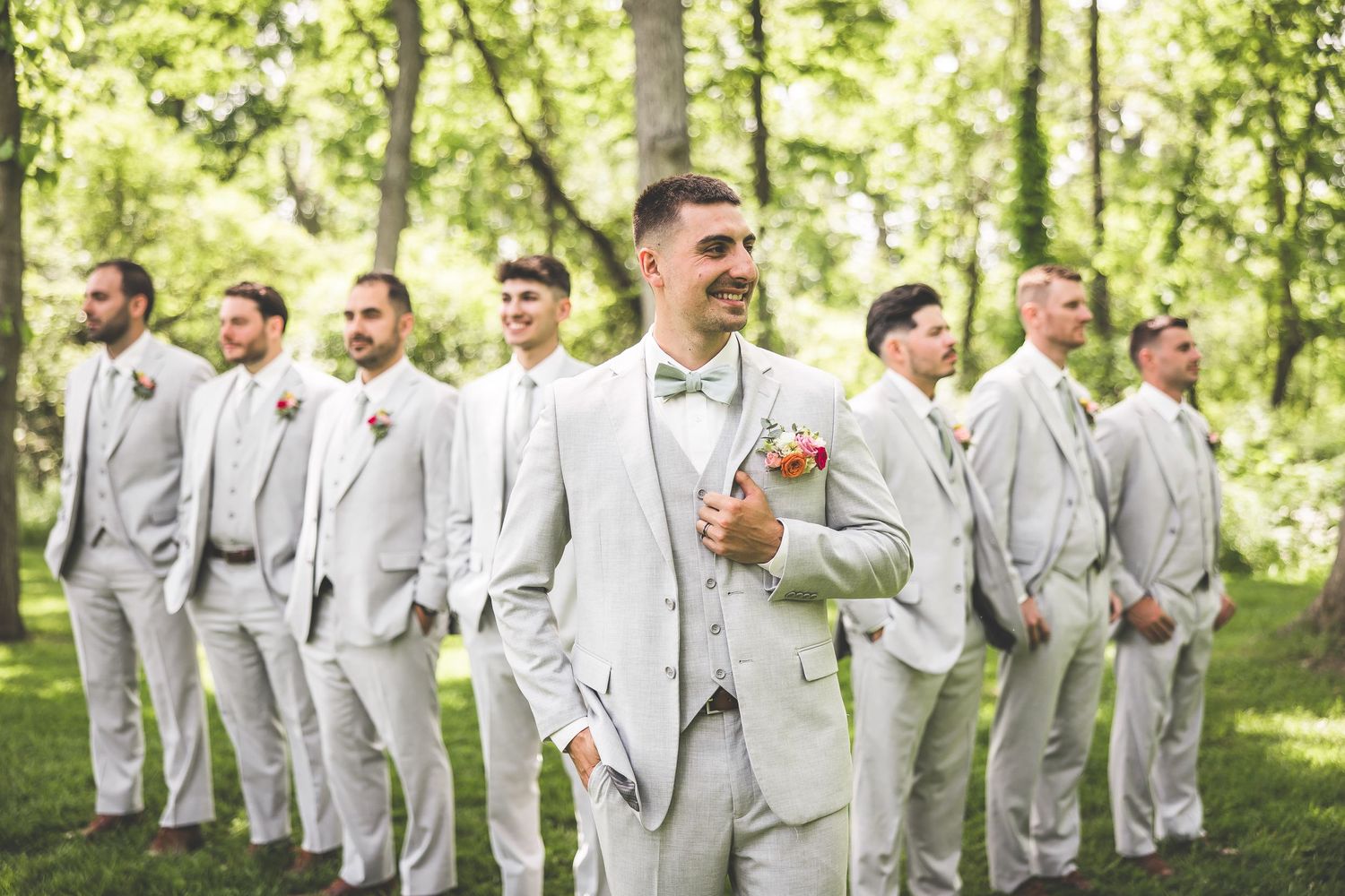 Groomsmen in light gray suits pose in a line outdoors.