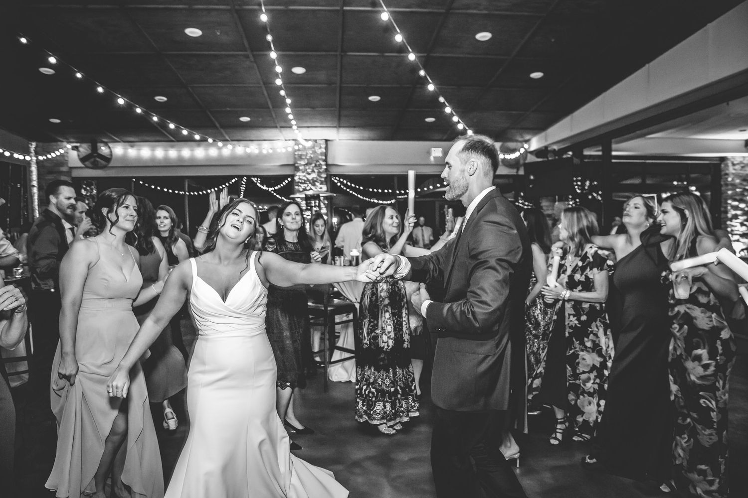 Black and white photo of wedding guests dancing under string lights at an indoor reception.