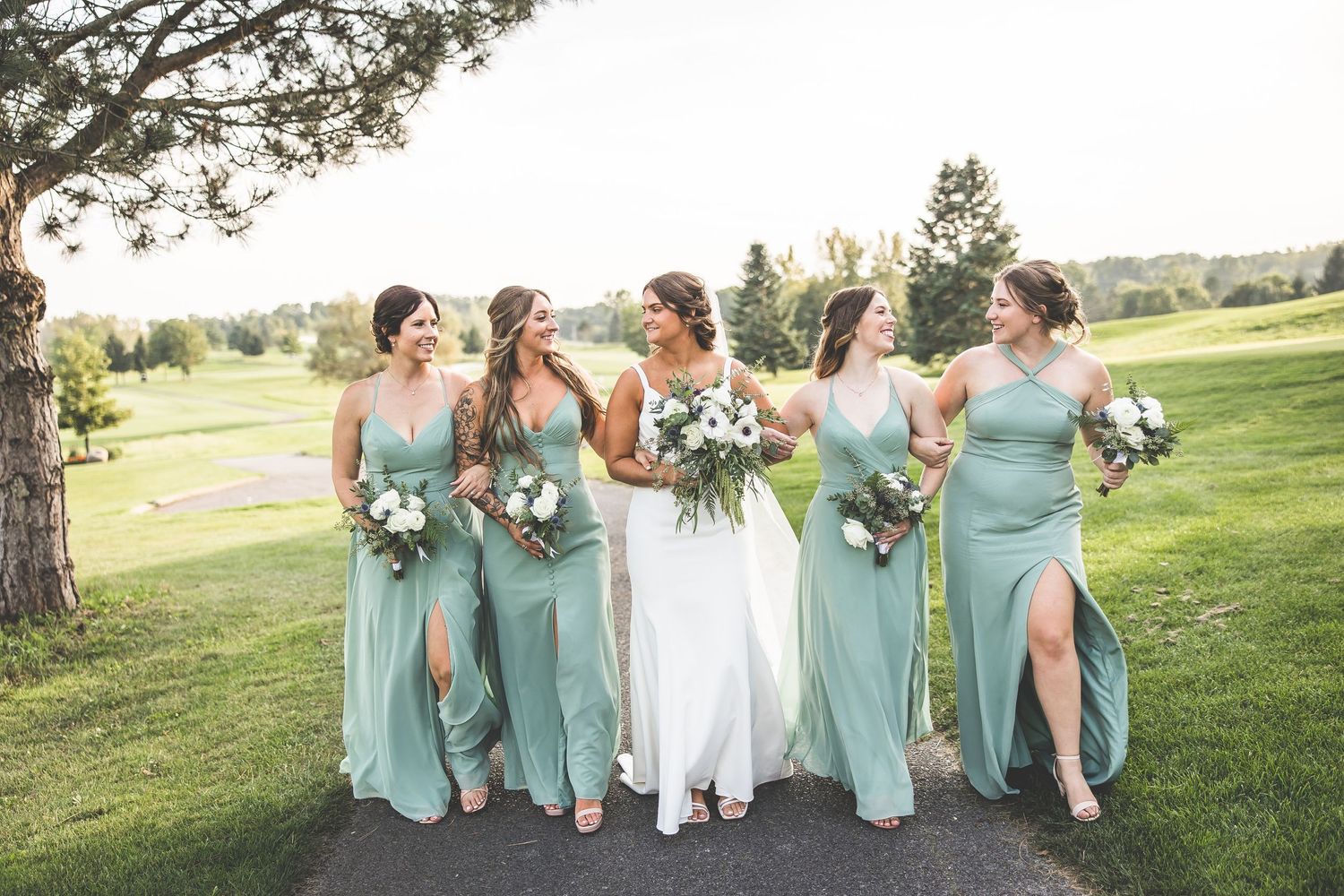Bride and bridesmaids in mint green dresses laugh together on golf course path.