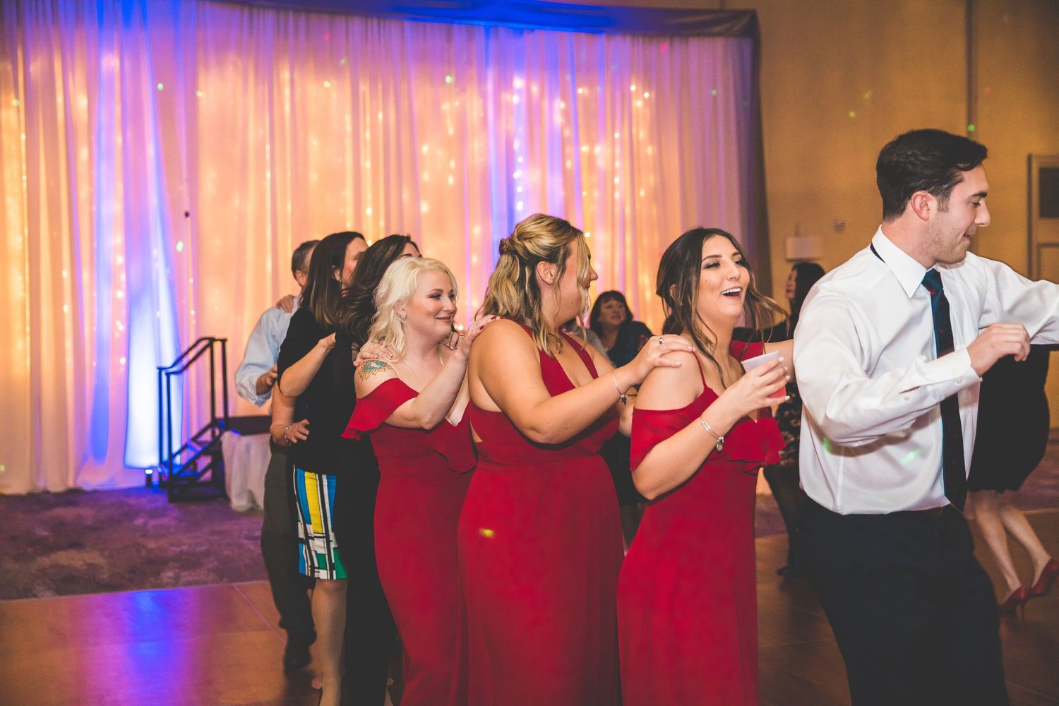 Wedding party members in red dresses and formal attire dancing at reception with colorful lighting.