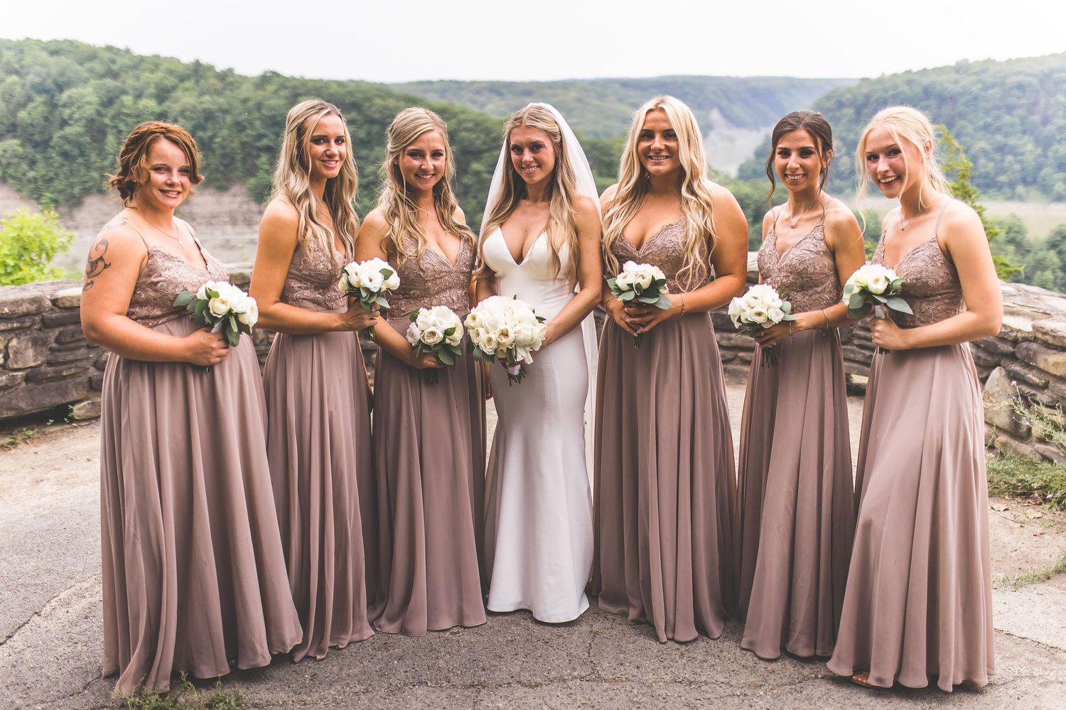 Wedding party in mauve bridesmaid dresses and black suits pose together on stone terrace overlooking mountains.