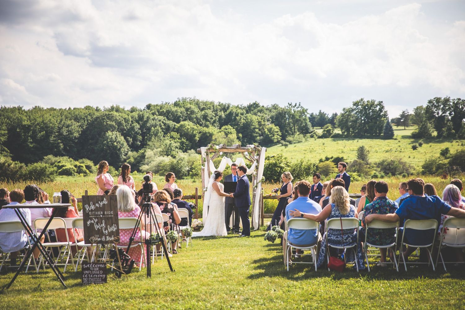 Row of white chairs set up on green grass for outdoor wedding ceremony with guests arriving.