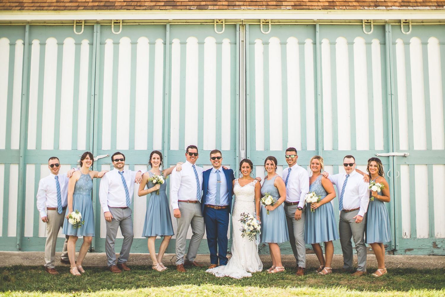Wedding party poses against a rustic barn wall in coordinated blue and white attire.