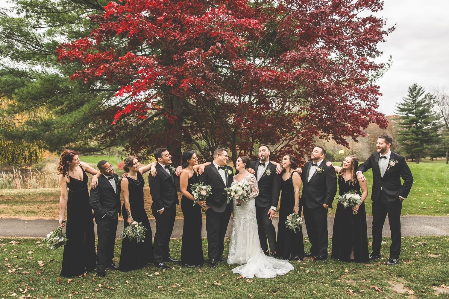 Large wedding party group photo with dramatic red maple tree and autumn foliage.