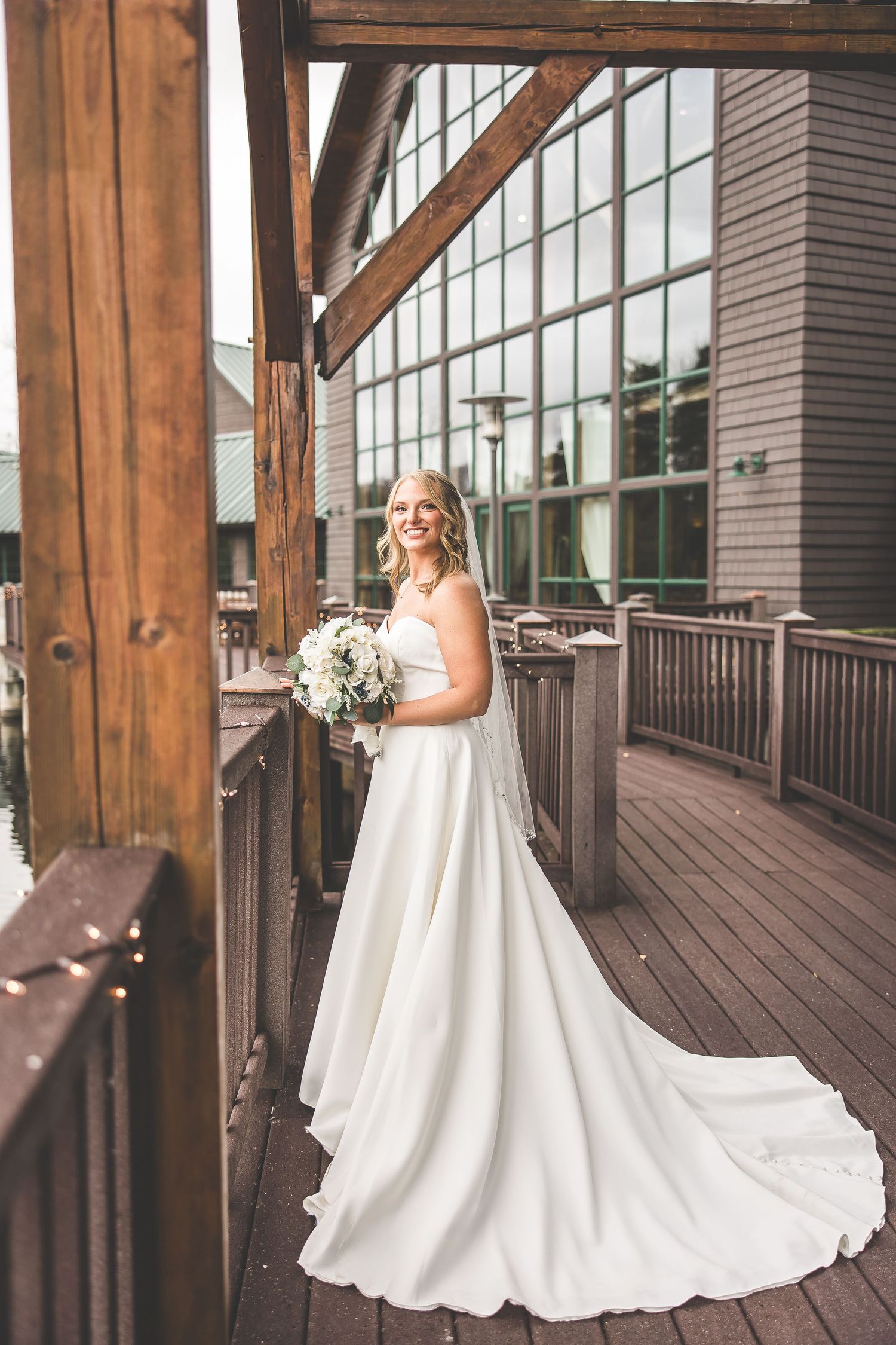 Elegant bride in flowing white gown stands on wooden deck of modern venue.