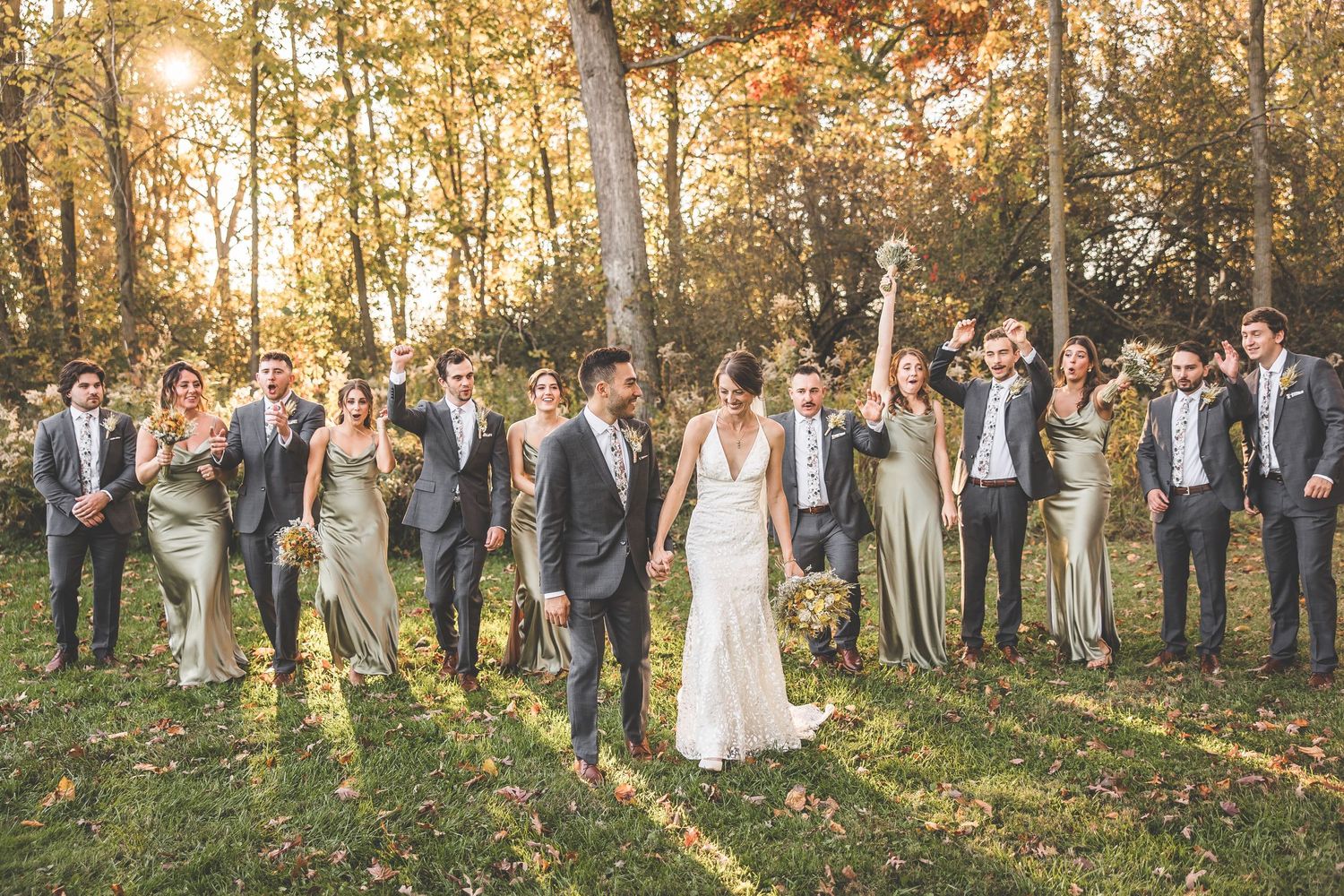 Large wedding party group photo in wooded setting with warm autumn lighting.