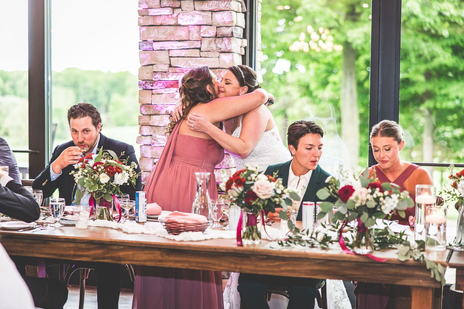 Long wooden table with floral centerpieces set for wedding reception by bright windows.