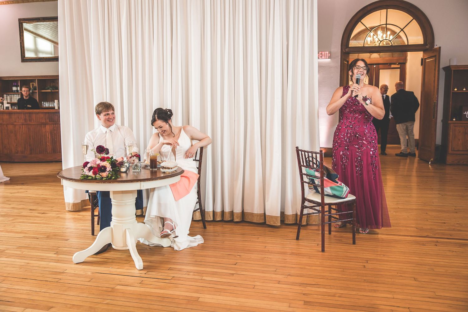 Wedding reception venue features hardwood floors, white drapes, and round dining tables.