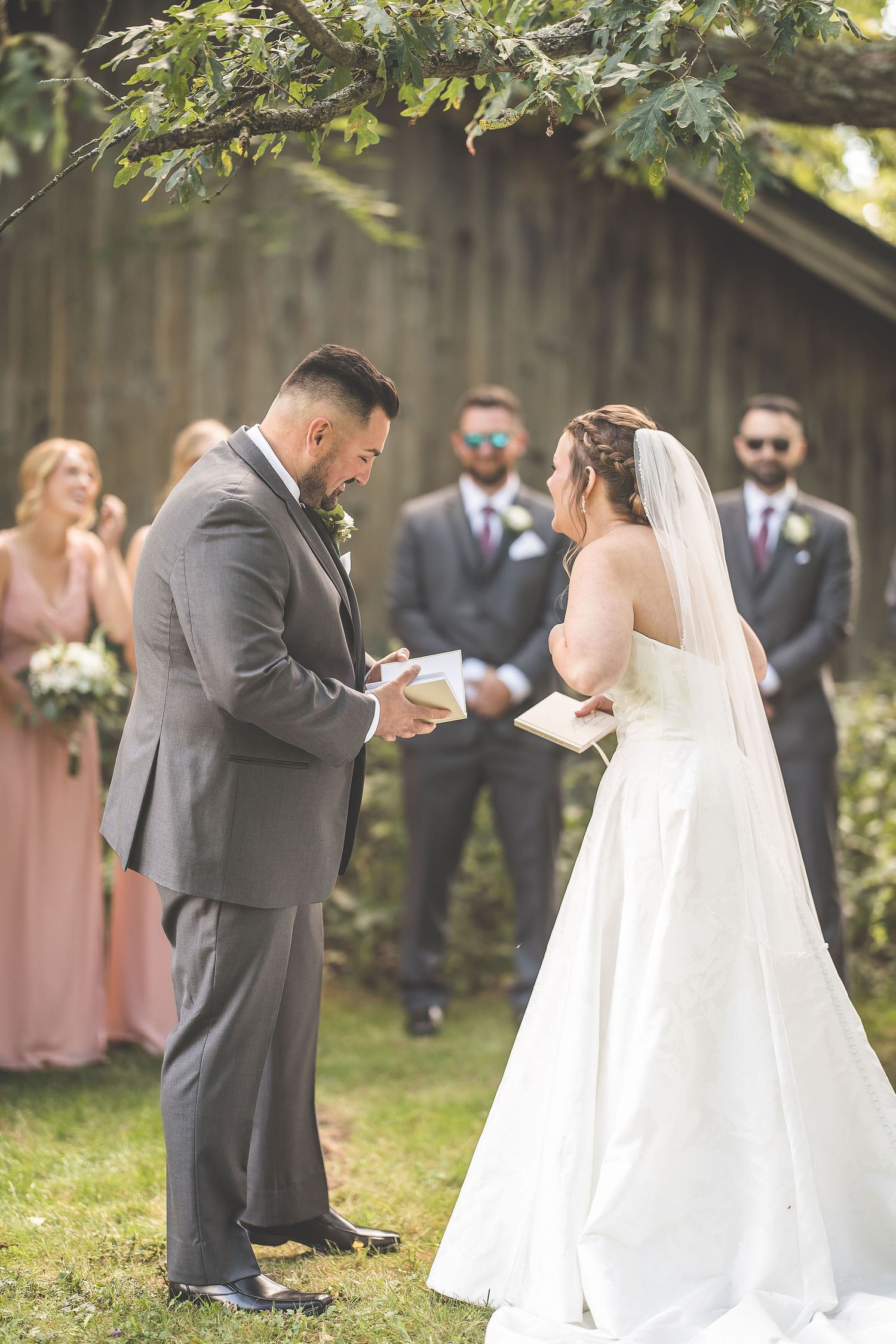 Wedding ceremony in progress with bridal party and guests watching under shaded trees.
