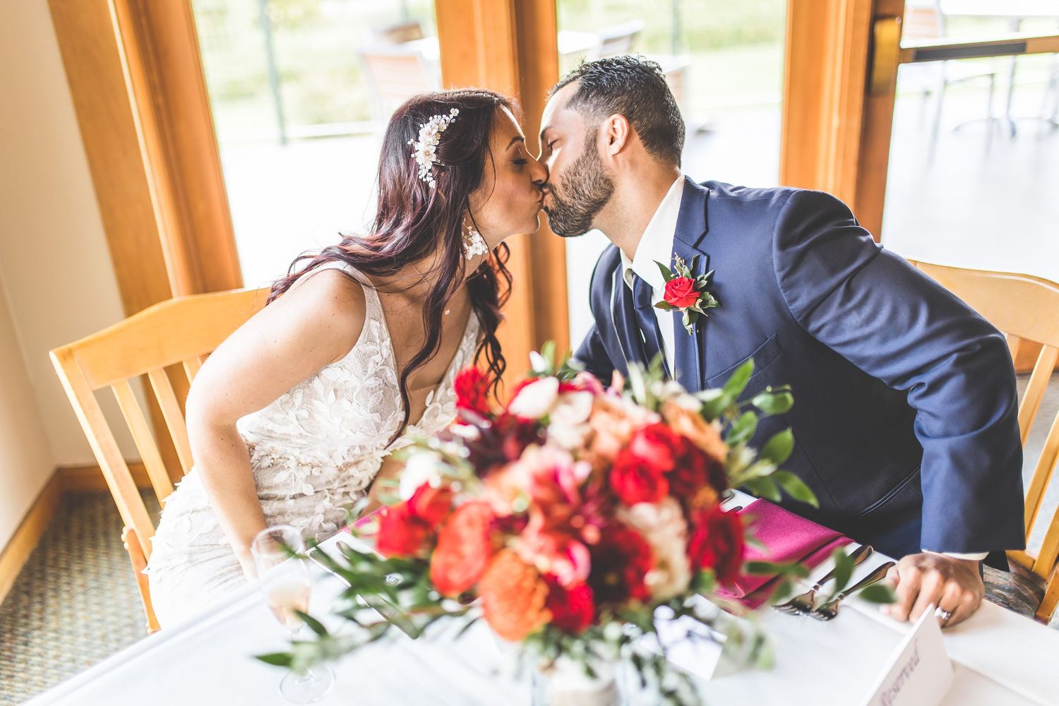 Intimate wedding reception scene with couple at dining table decorated with red flowers.
