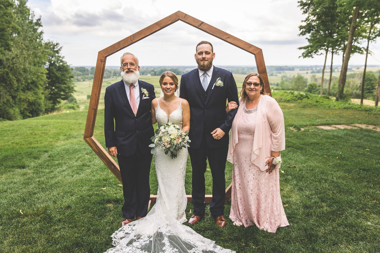 Wedding party poses under geometric wooden arch with rolling hills in background.