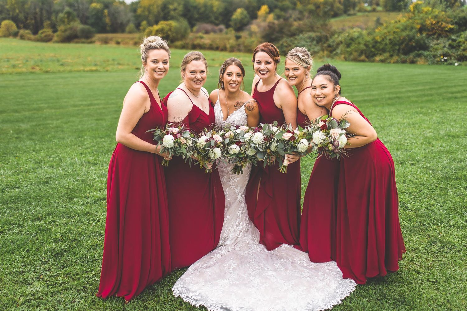 Wedding party poses outdoors with bridesmaids in red dresses on grassy hillside.