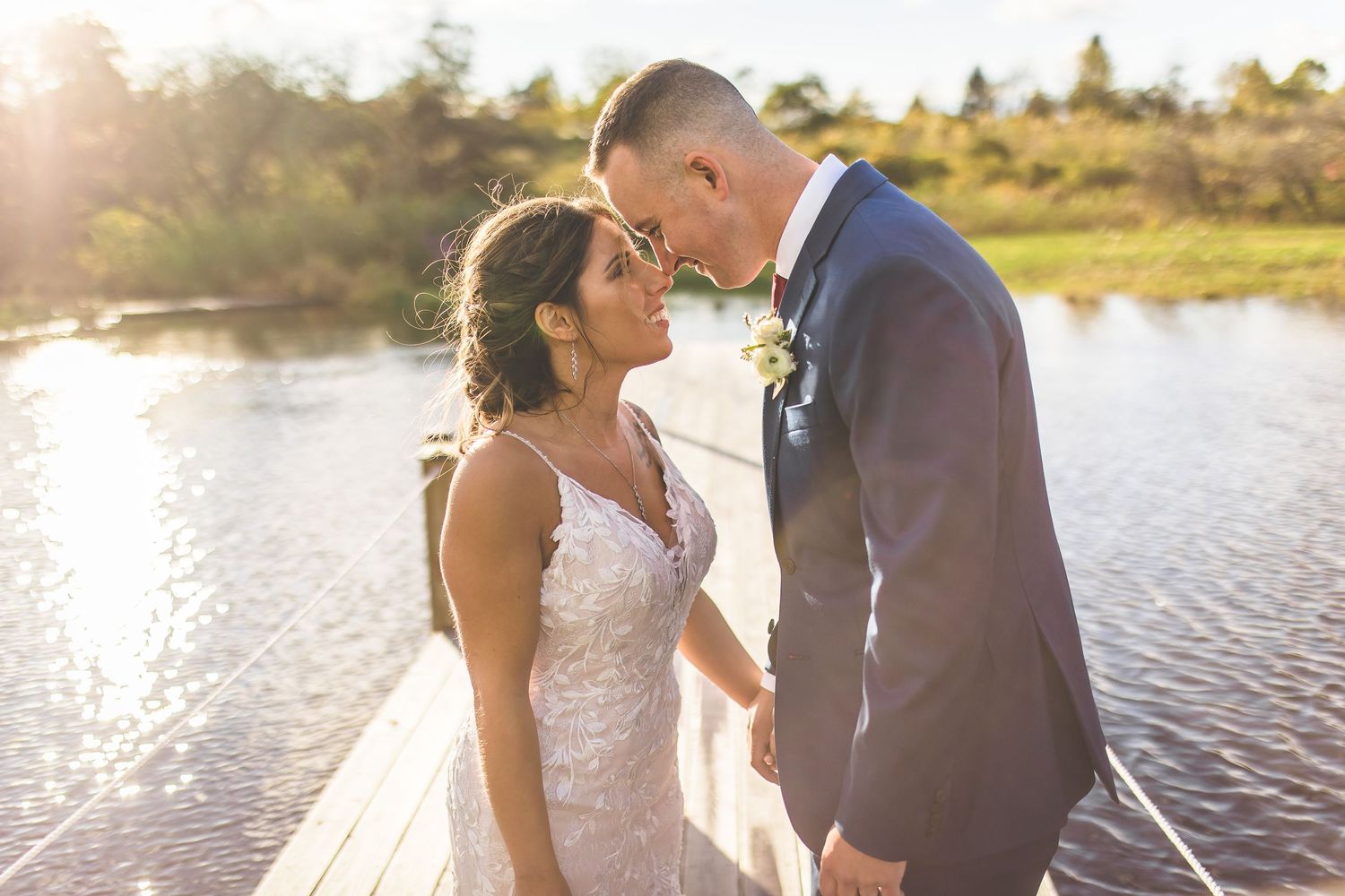 Bride in lace dress and groom in navy suit share intimate moment by the water.