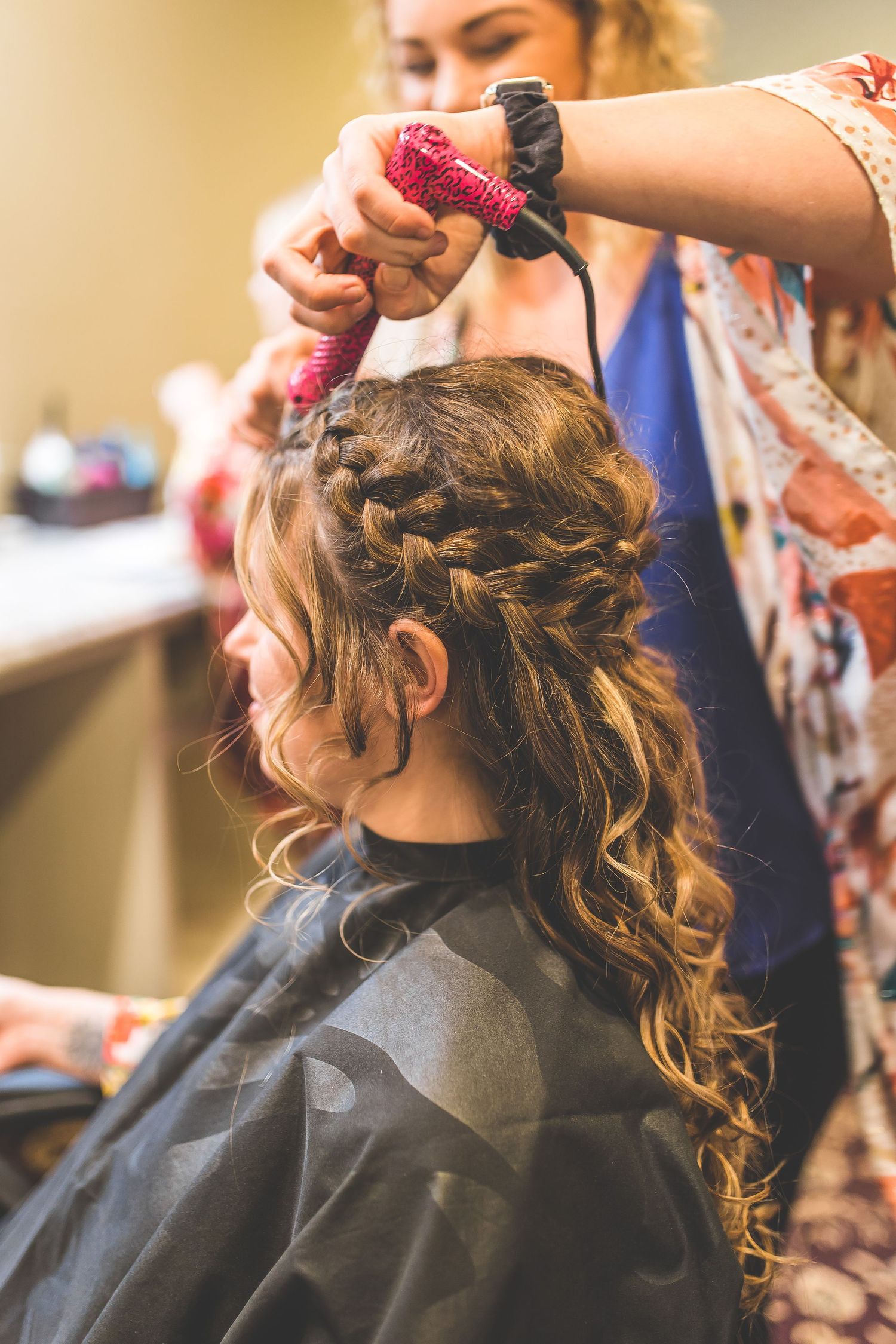 Hairstylist using a pink styling tool to create an intricate braided hairstyle with curls.