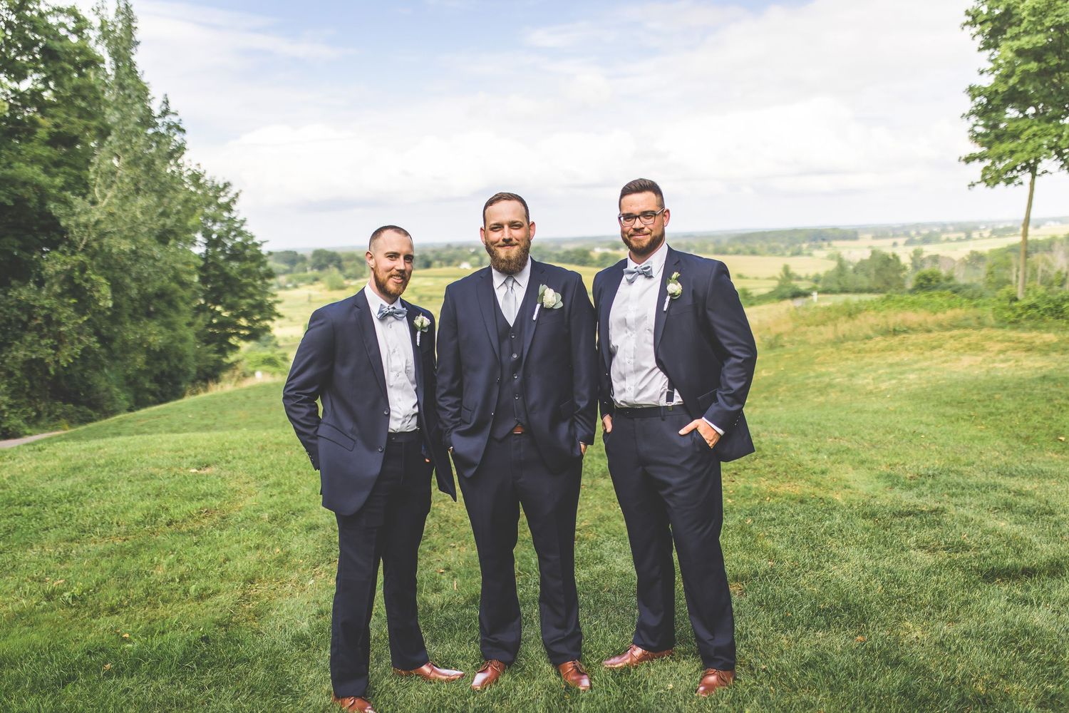 Groomsmen in dark suits pose casually on green hillside during outdoor wedding.