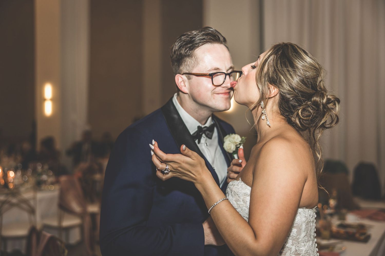 Candid wedding moment captured as couple shares a tender kiss in a warmly lit reception venue.