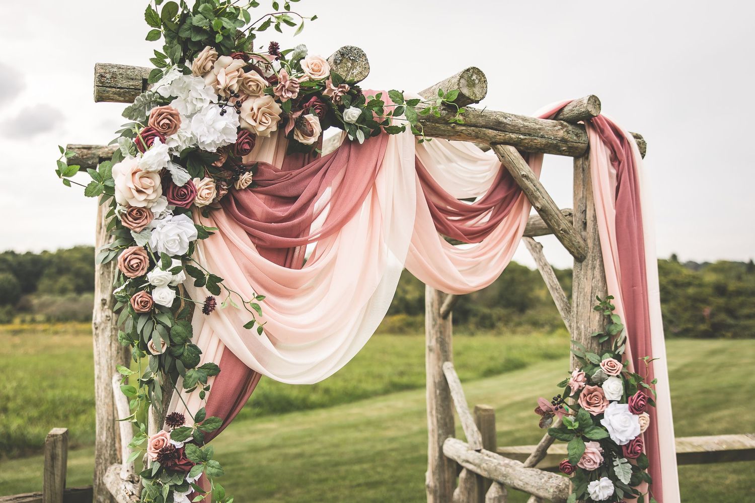 Rustic wooden wedding arch decorated with pink fabric draping and floral arrangements in an outdoor setting.