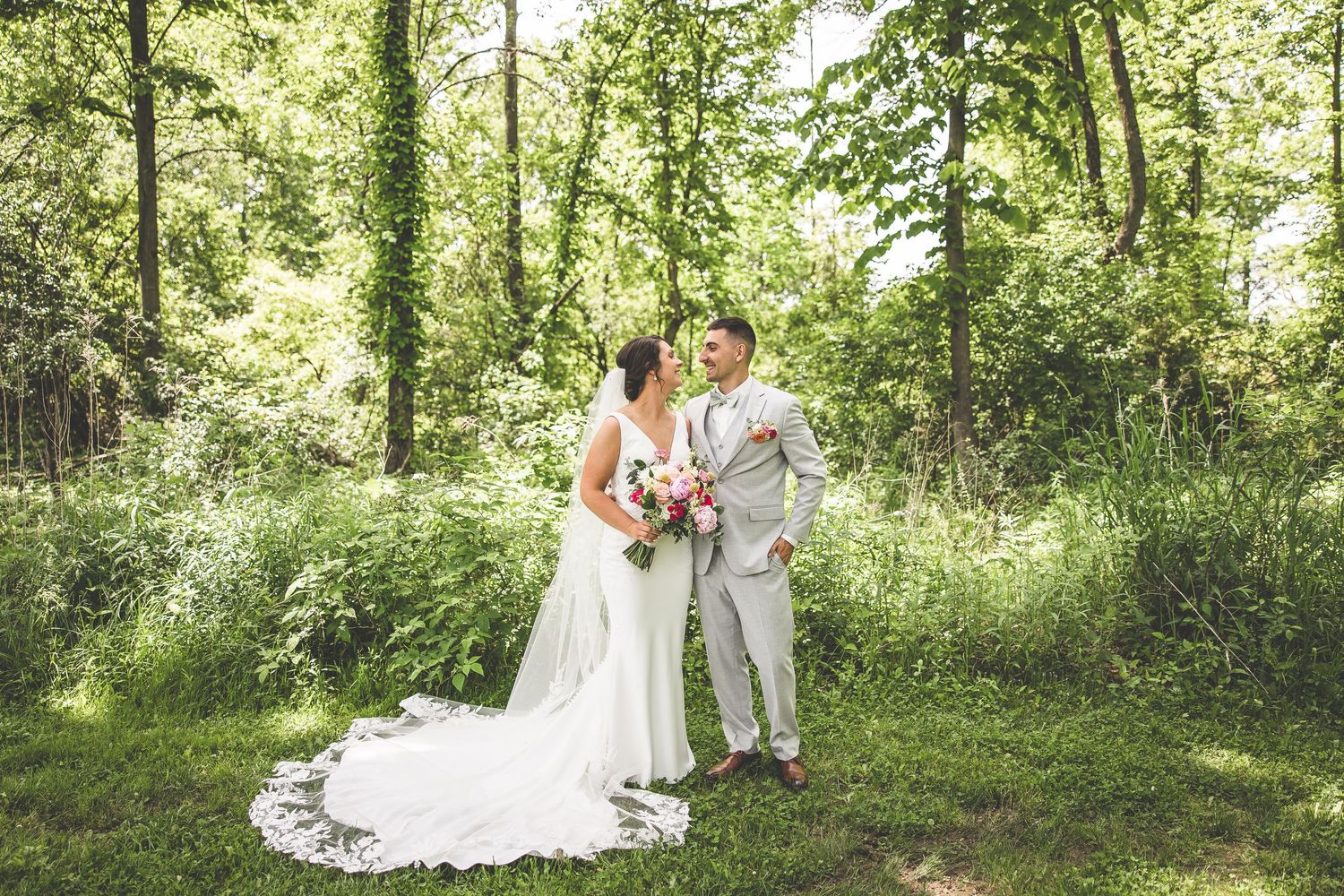 A couple in wedding attire shares an intimate moment in a sunlit forest clearing.
