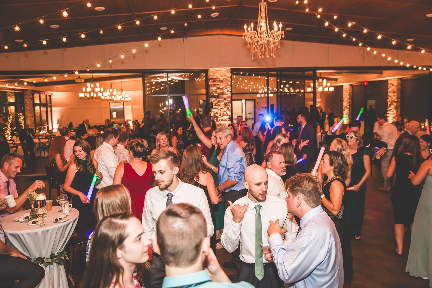 Packed dance floor at wedding reception with guests celebrating under string lights and chandeliers.