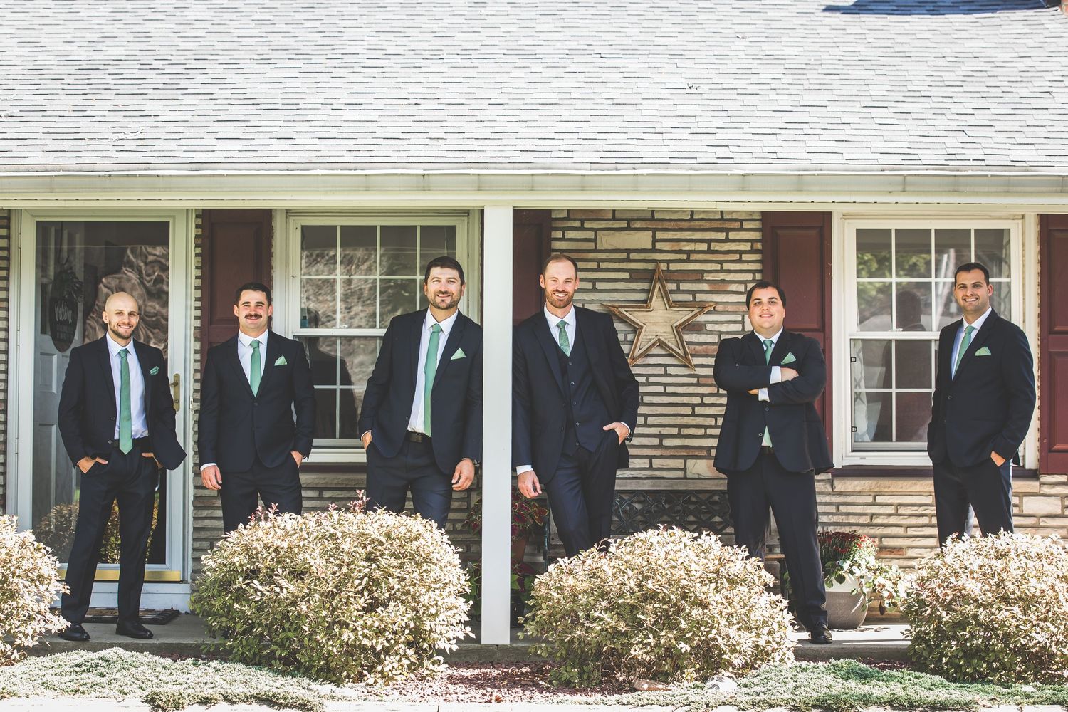 Wedding party in formal attire gathered outside traditional stone house.