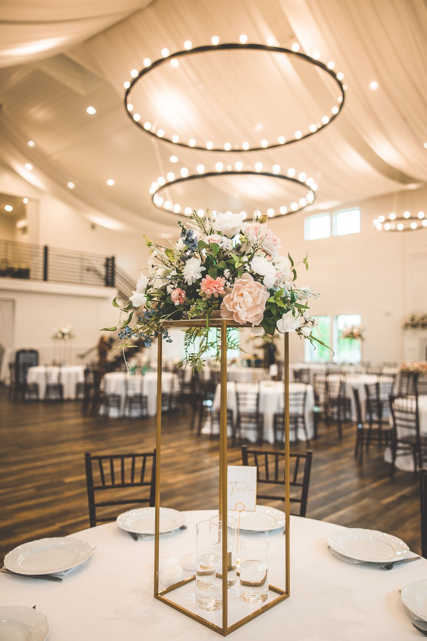 A modern wedding reception table with tall floral centerpiece under circular lighting fixtures.