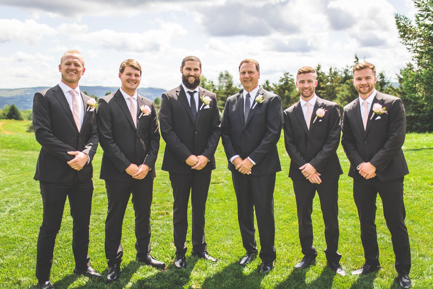 Groomsmen in black suits with pink accents pose together on a sunny outdoor lawn.