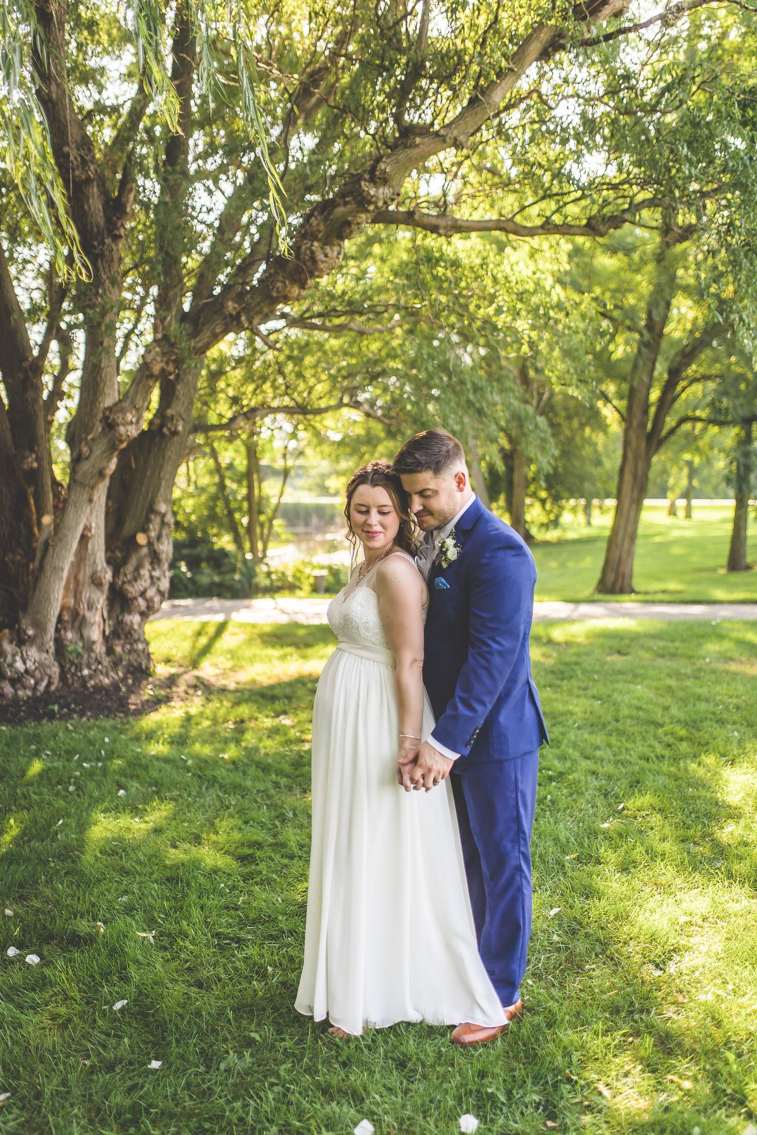 A romantic outdoor wedding portrait featuring a couple in a white dress and blue suit under sunlit trees.