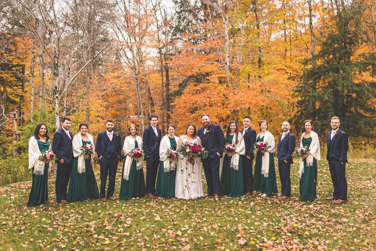 Large wedding party group photo with autumn trees and fallen leaves in background.