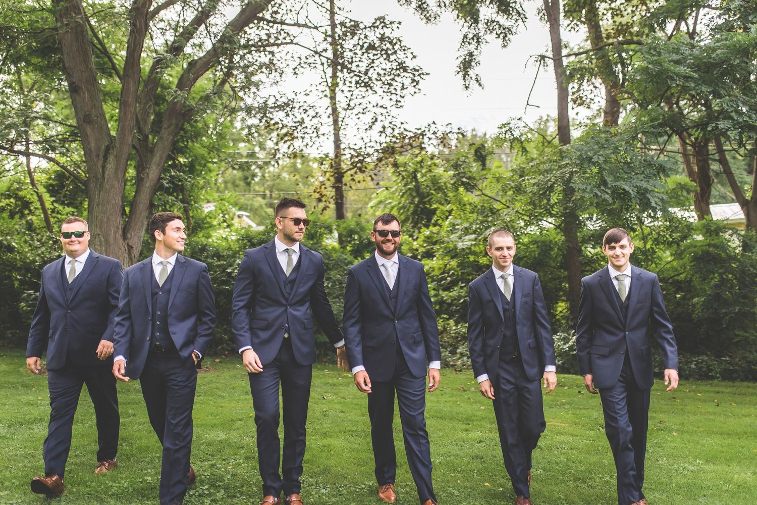 Wedding party in navy suits walking together through a lush green garden.