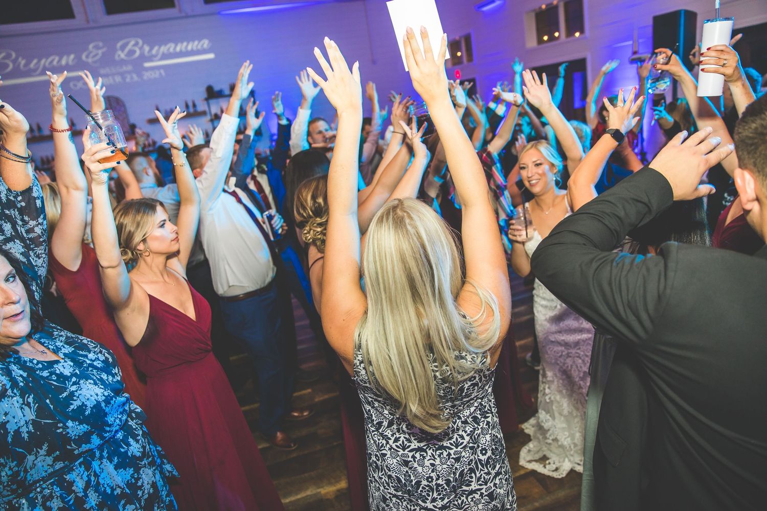 Guests with raised hands dancing and celebrating at a wedding reception with colorful lighting.