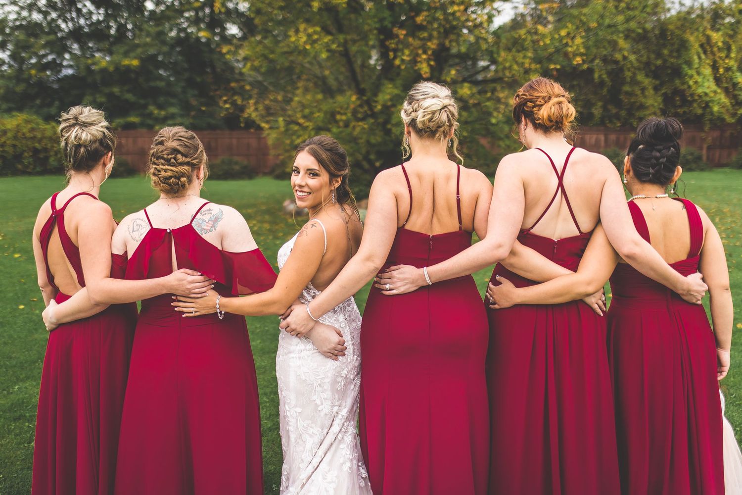 Wedding party embracing in a line outdoors showing backs of red bridesmaid dresses.