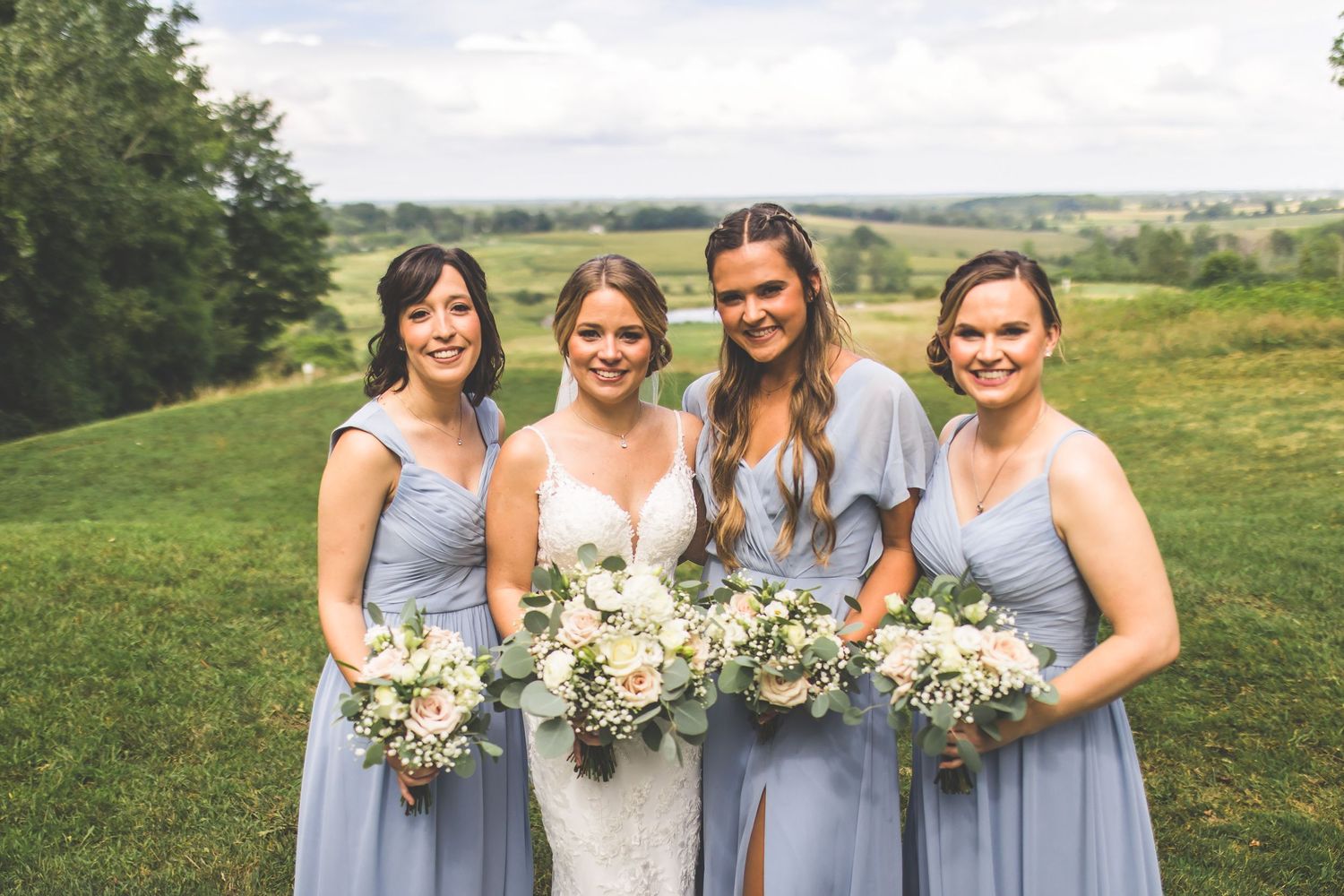 Wedding party stands together outdoors with bridesmaids in pale blue dresses holding white bouquets.