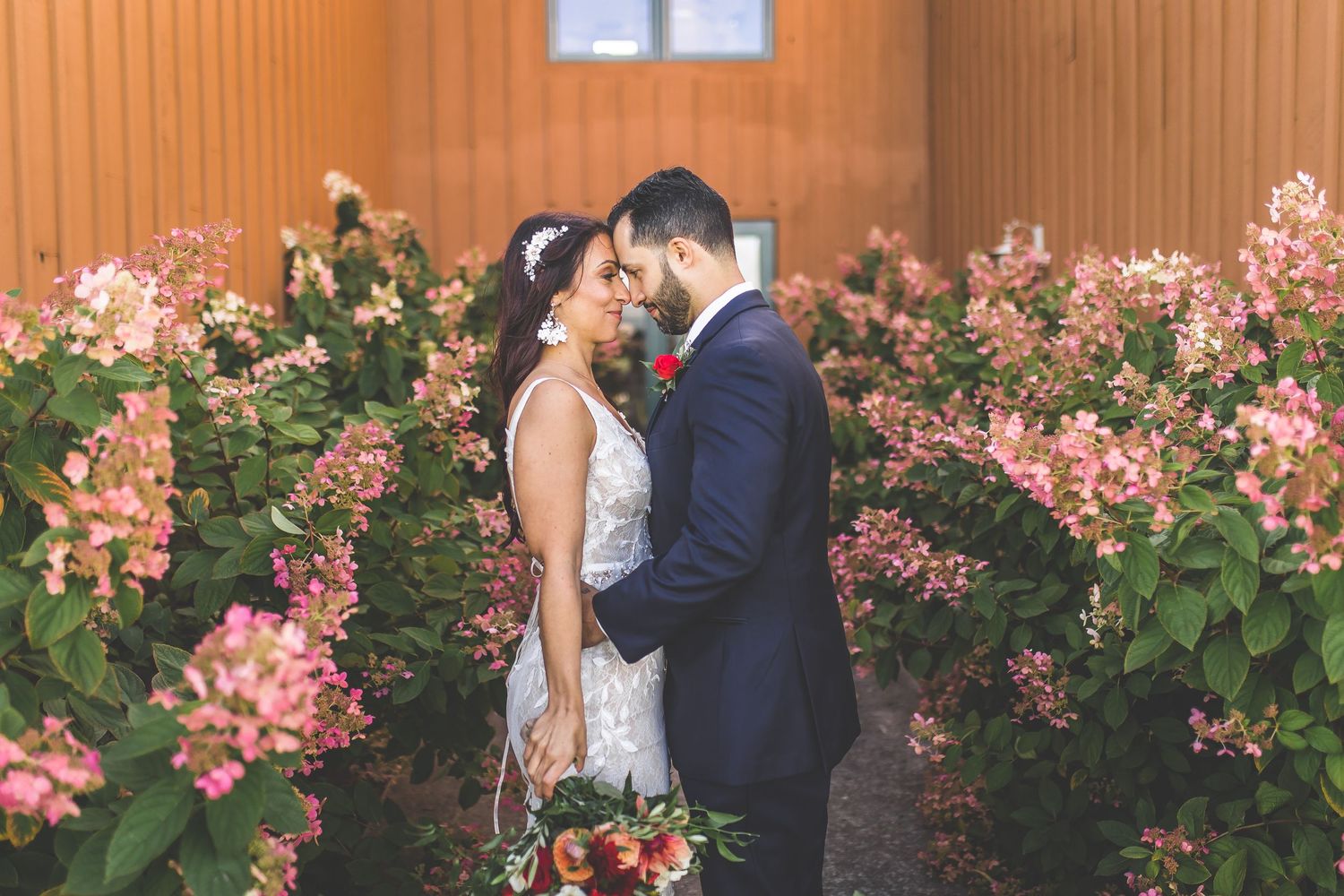Wedding party poses in front of pink flowering bushes at outdoor venue.
