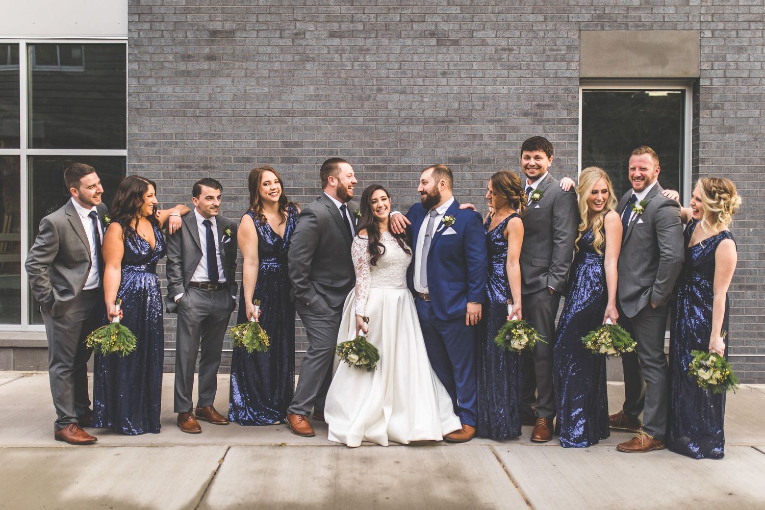 Wedding group photo with bridal party in navy dresses and gray suits against urban backdrop.