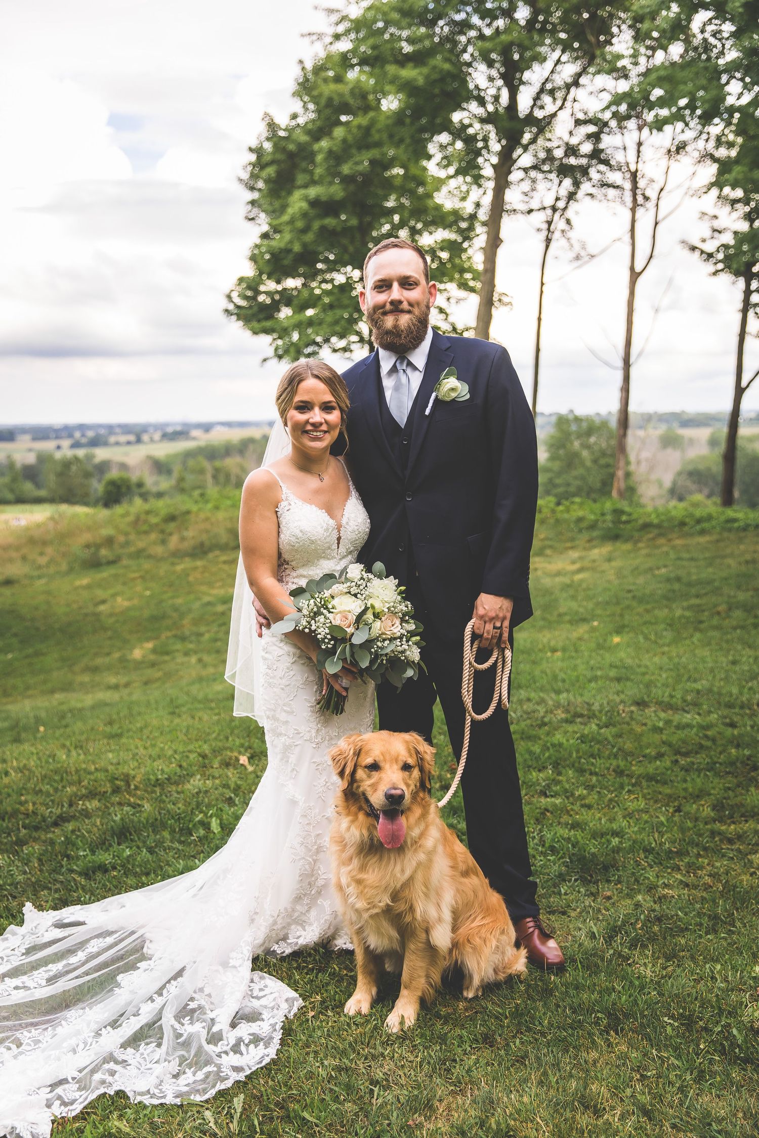 Wedding portrait with couple and their golden retriever on hilltop overlooking scenic landscape.