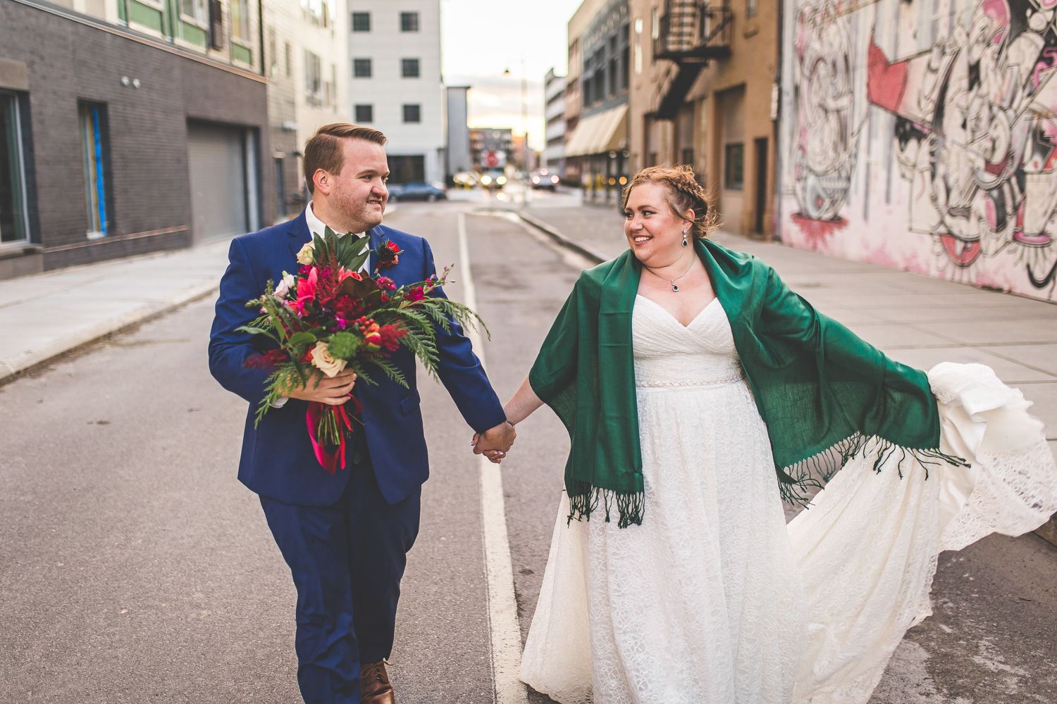 Couple walks hand in hand down urban street, bride in white dress with emerald shawl.