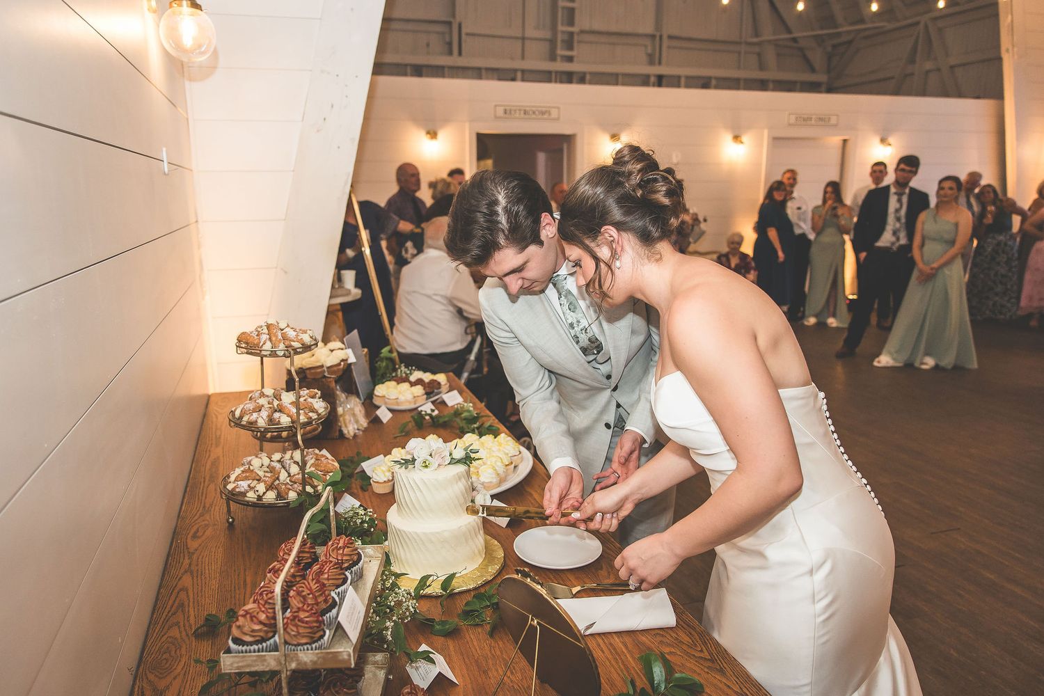 Wedding reception guests gather around tables with decorative elements in an elegant white venue.