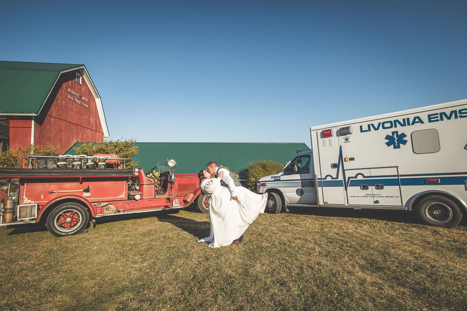 Panoramic shot of emergency vehicles and wedding party gathered on a hillside near a red barn.
