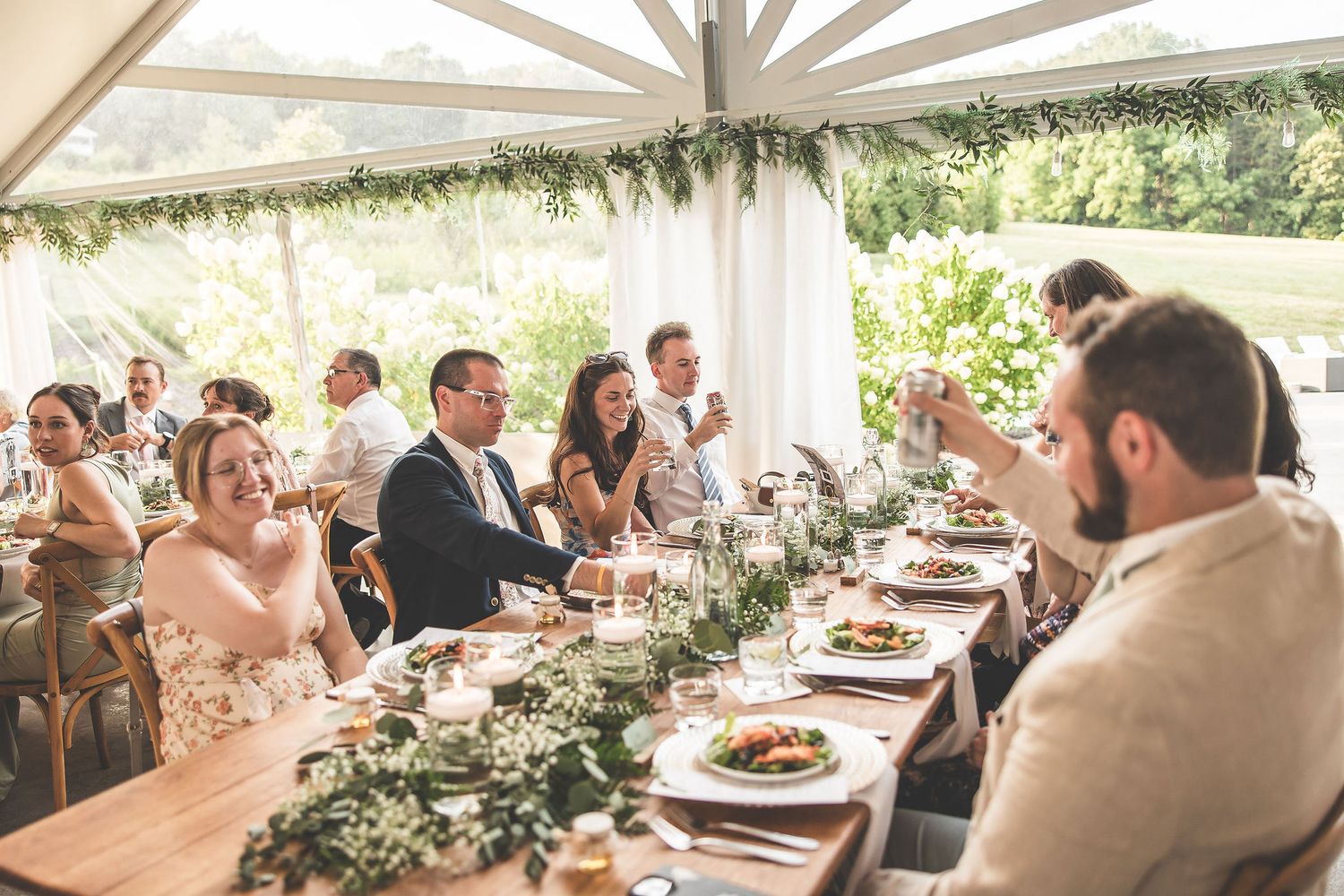 Wedding guests enjoy an outdoor reception under a white tent with string lights and natural greenery.