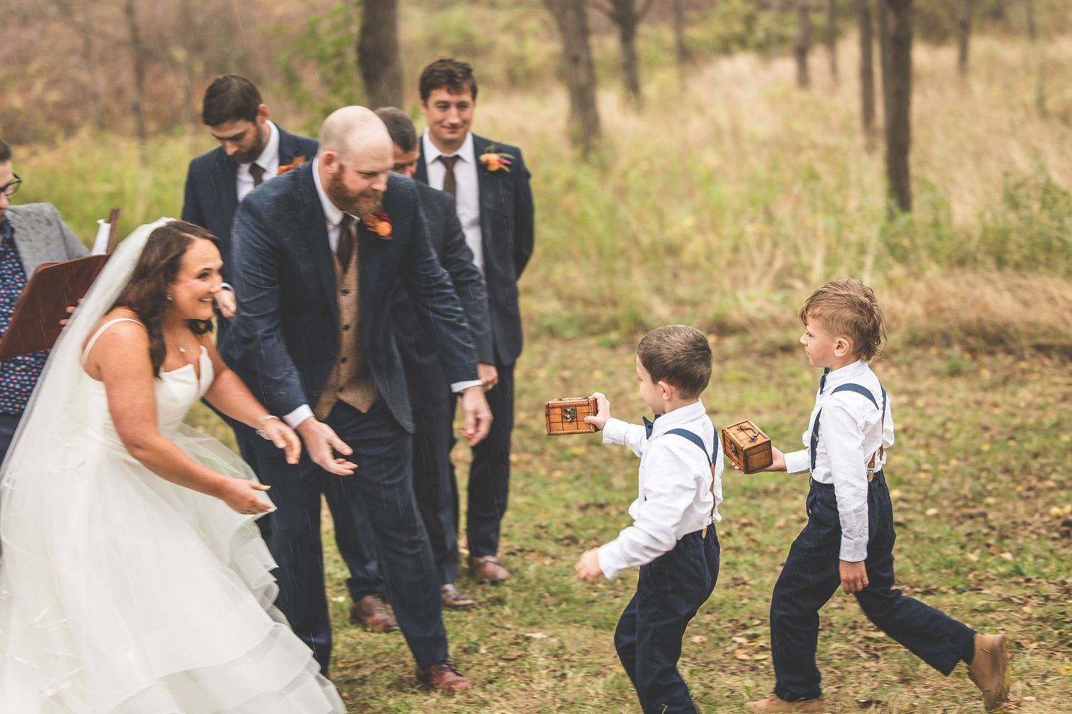 Young ring bearers in suspenders approach the wedding party during outdoor ceremony.