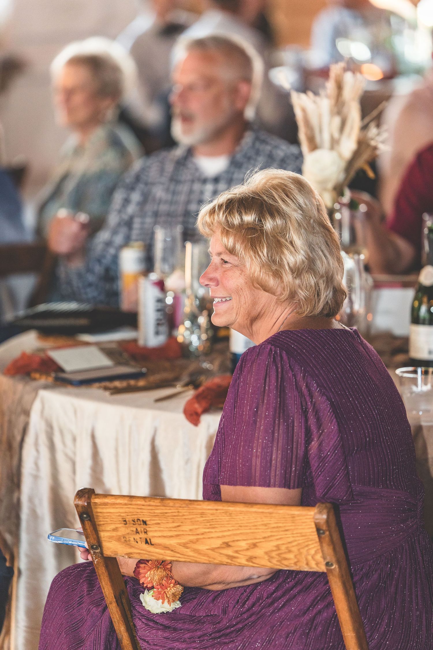 Wedding guests in purple attire enjoy the reception from wooden chairs.