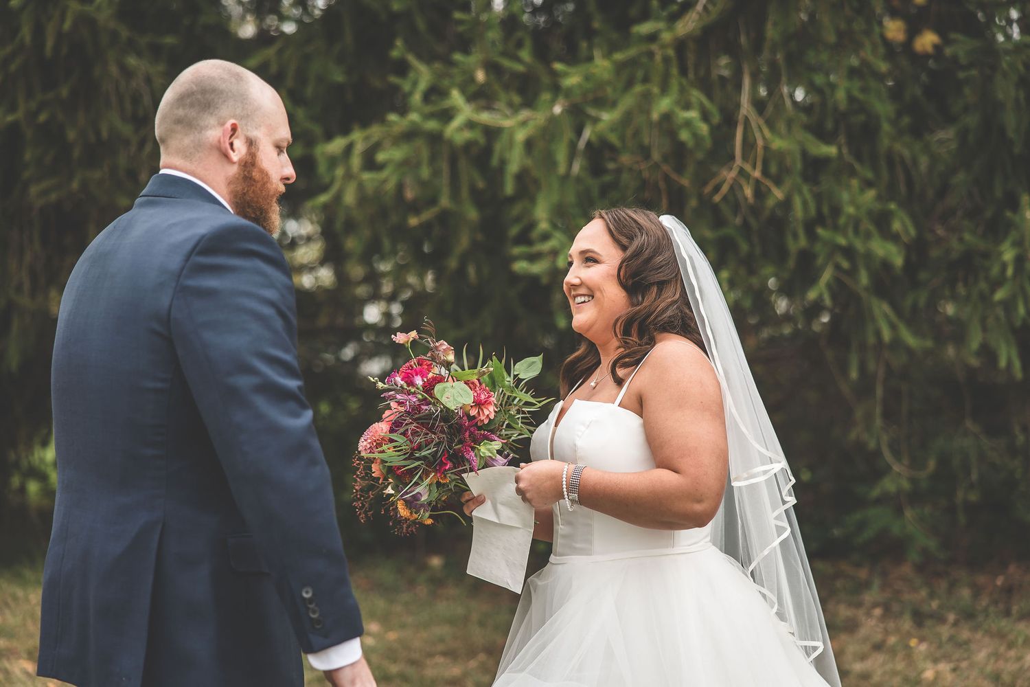 Wedding couple shares candid moments during their first look photoshoot against evergreen trees.