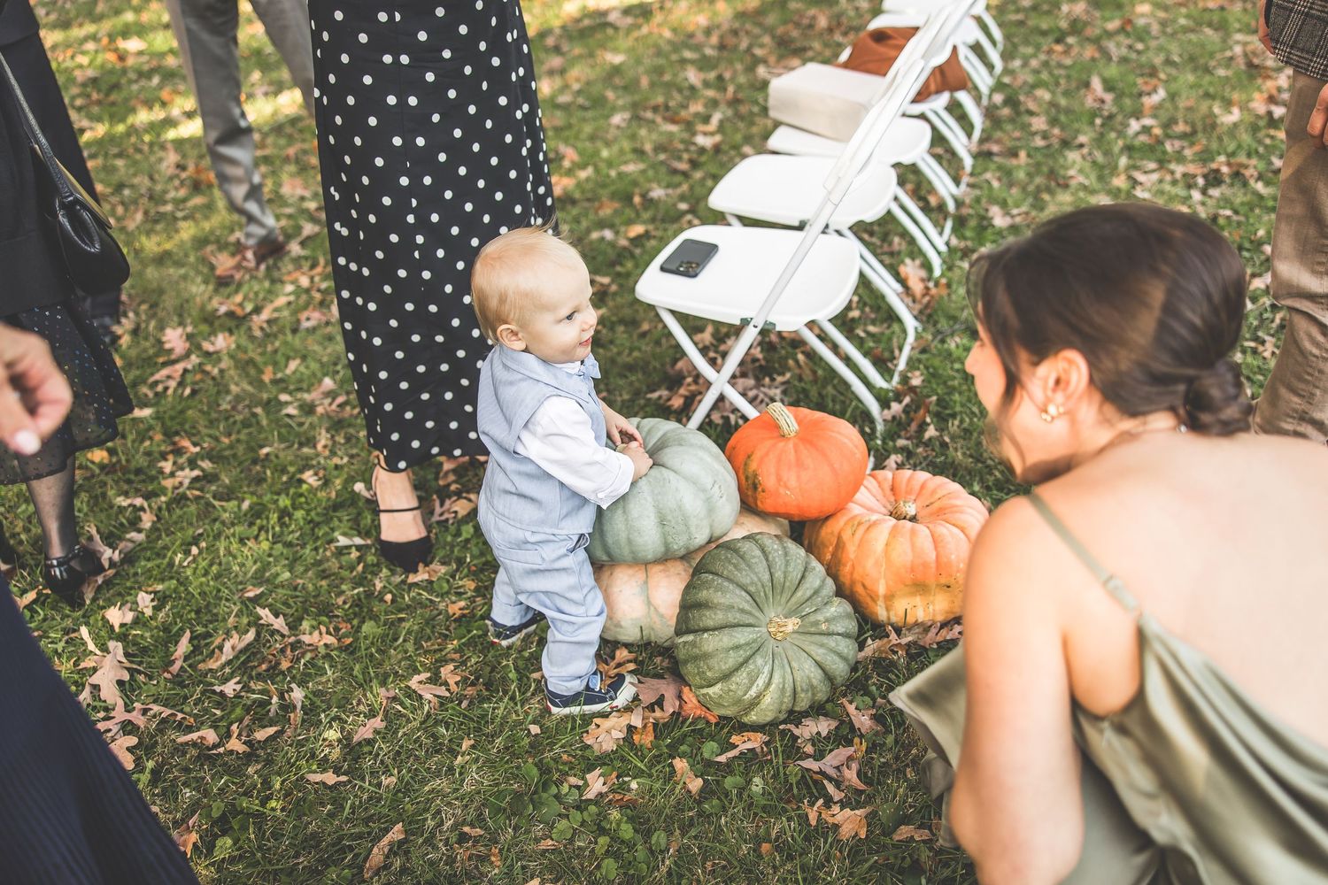 Wedding guests interact with pumpkin decorations on grass during an outdoor fall celebration.