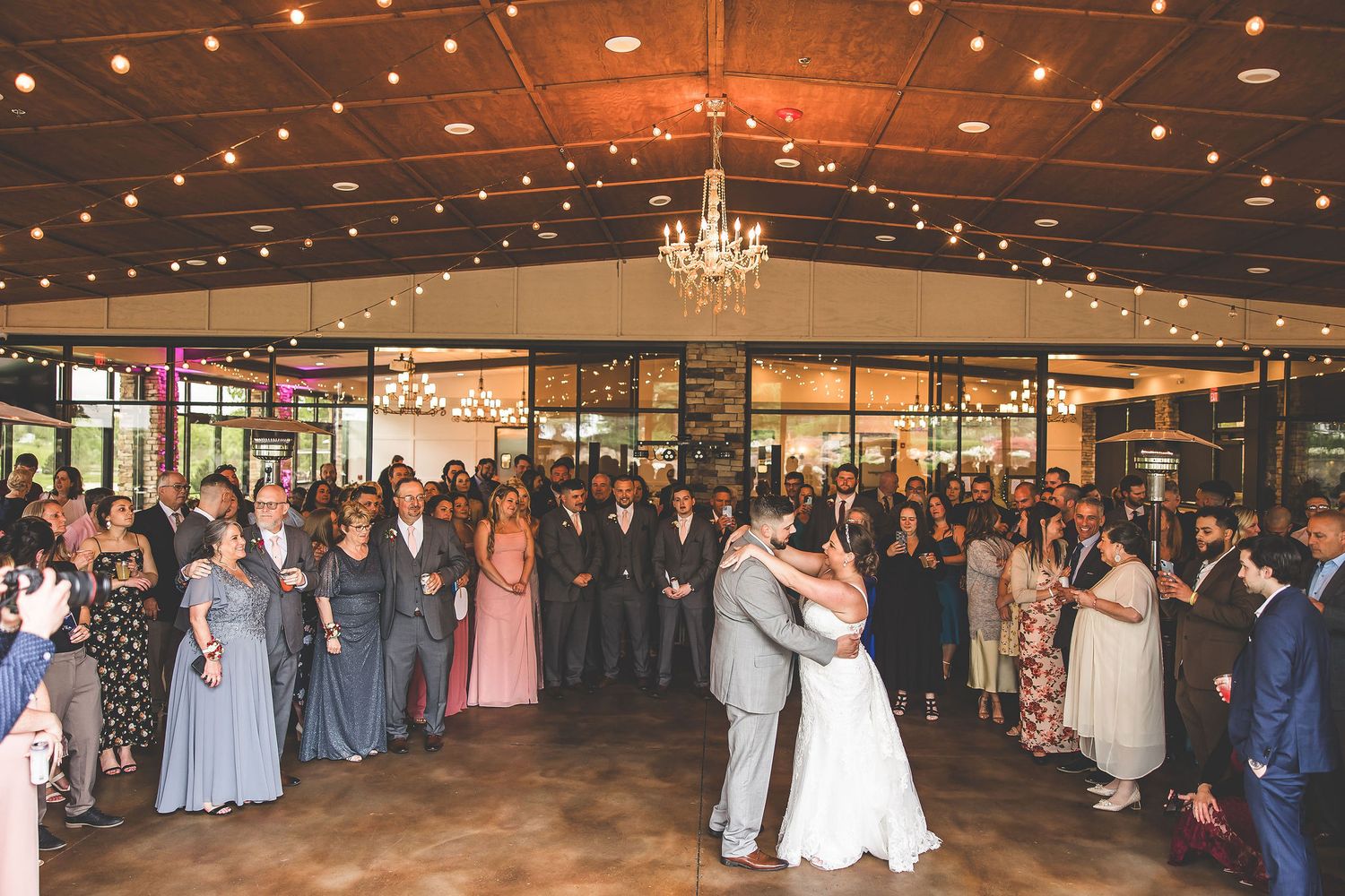 Wedding reception venue decorated with string lights and chandeliers as guests celebrate.